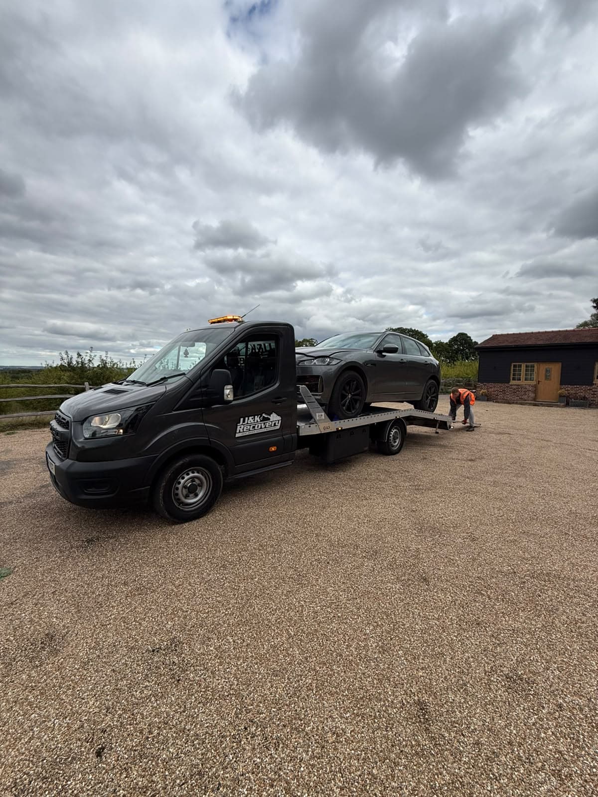 J&K Recovery truck with a grey SUV on the flatbed while an operator secures it at a countryside property